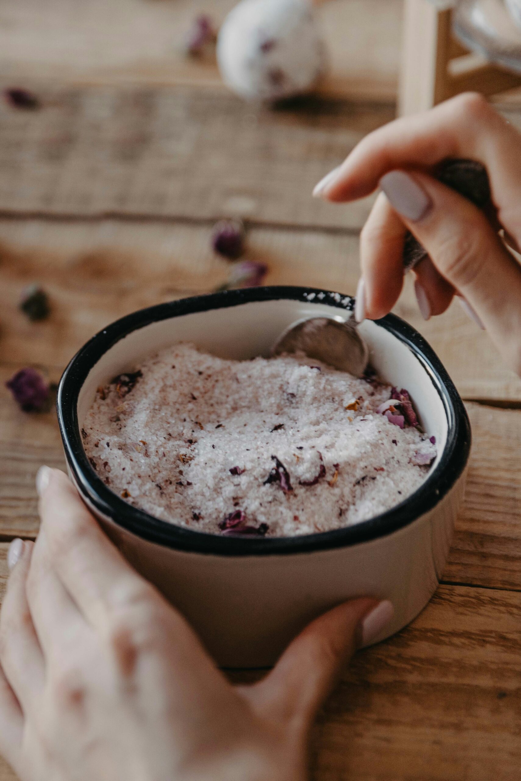 Close-up of a hand holding a spoon above a bowl of pink Himalayan salt mixed with dried rose petals, set on a wooden table. The image captures a serene and natural aesthetic, ideal for wellness and relaxation themes.