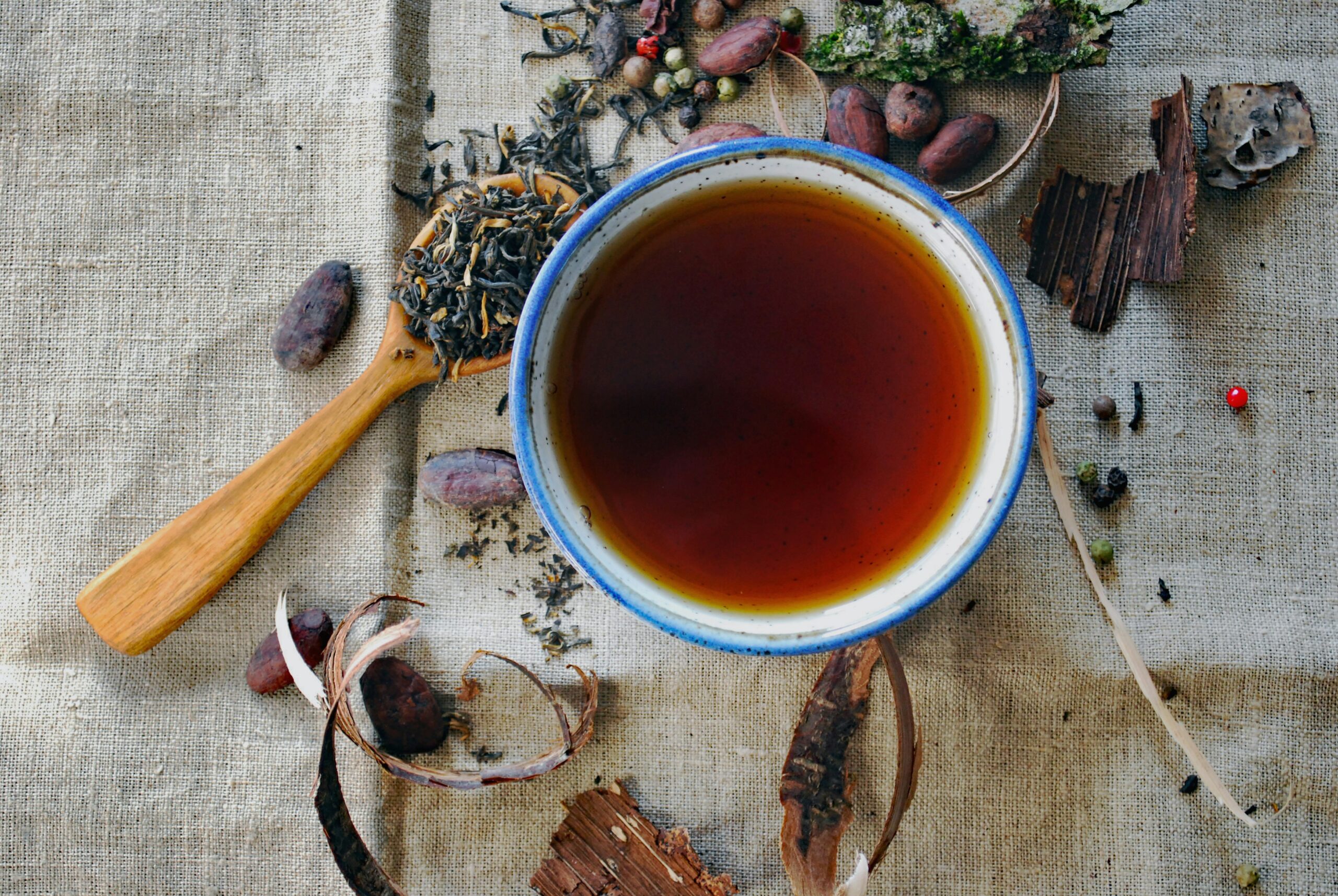 A top-down view of a blue-rimmed bowl filled with dark brown tea, surrounded by loose tea leaves, cocoa beans, and natural textures on a burlap background. The image captures the essence of tea preparation and the organic materials associated with it.