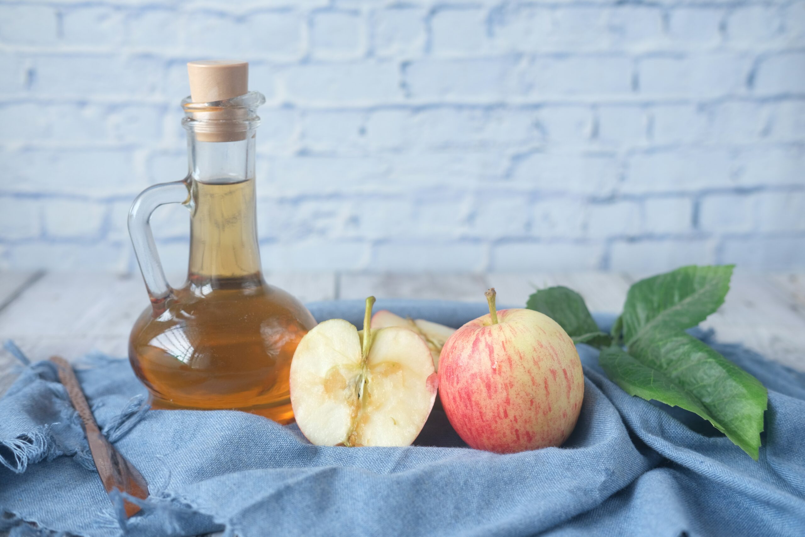 Glass bottle of apple cider vinegar next to whole and halved fresh apples on a blue cloth, with a wooden spoon in the background. Ideal for healthy cooking and natural remedies.