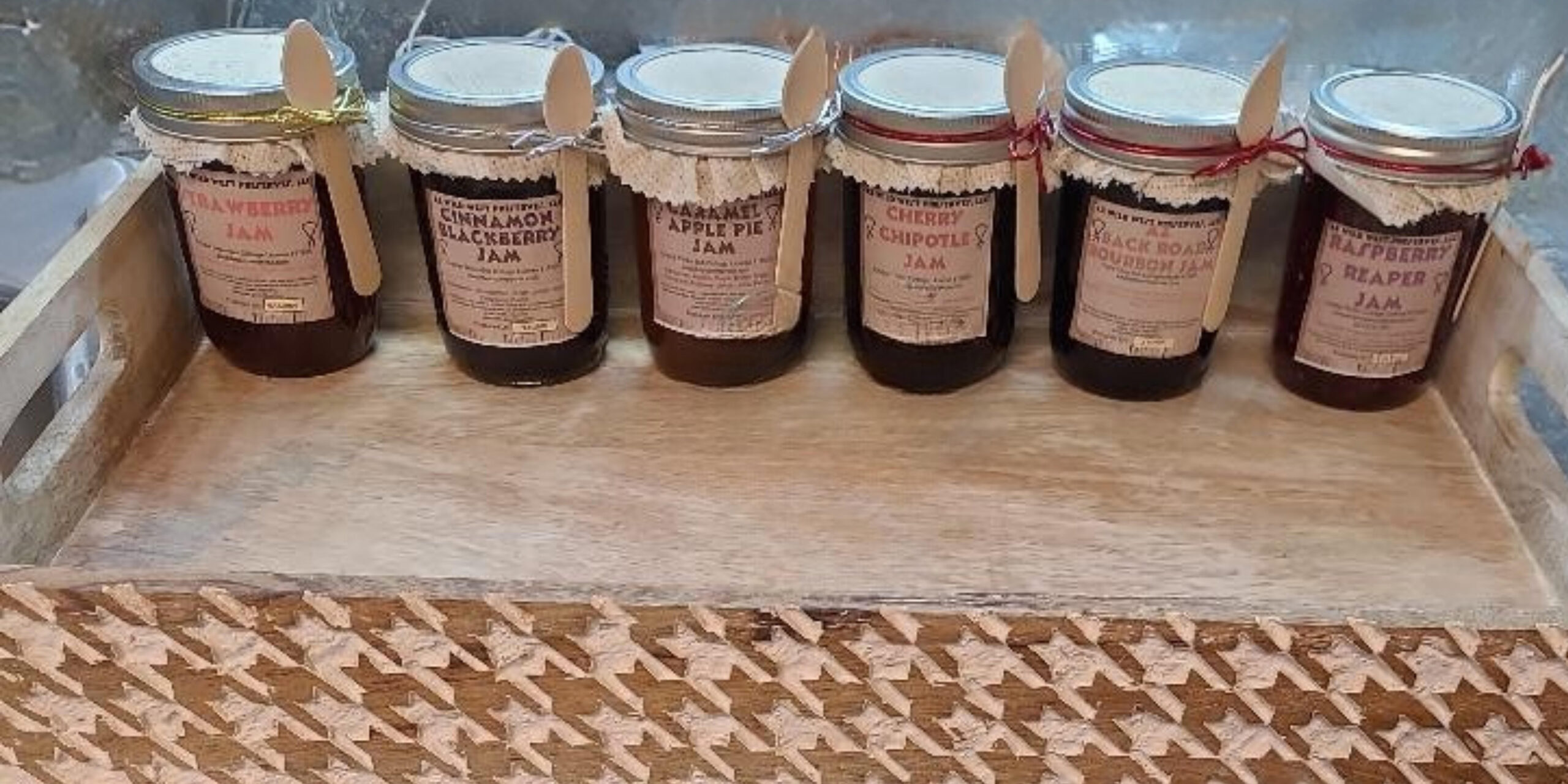 Assorted jars of gourmet jams displayed on a wooden tray, featuring flavors like Strawberry, Cinnamon Blackberry, Caramel Apple Pie, Cherry Chipotle, Black Road Bourbon, and Raspberry Reaper. Each jar is labeled and adorned with a decorative lid and small spoon.